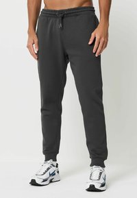 Dunkelgraue Jogginghose mit elastischem Bund und Kordelzug, ausgestattet mit enganliegenden Bündchen und glatter Stoffoberfläche. Getragen mit weißen Turnschuhen.