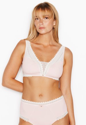 Donna con capelli biondi che indossa lingerie rosa chiaro con rifiniture in pizzo bianco, in piedi davanti a uno sfondo bianco semplice.