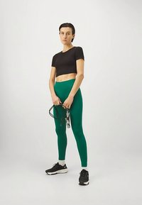 Fekete crop topot és zöld leggingset viselő nő, aki fekete és szürke ugrálókötéllel áll egy sima fehér háttér előtt.