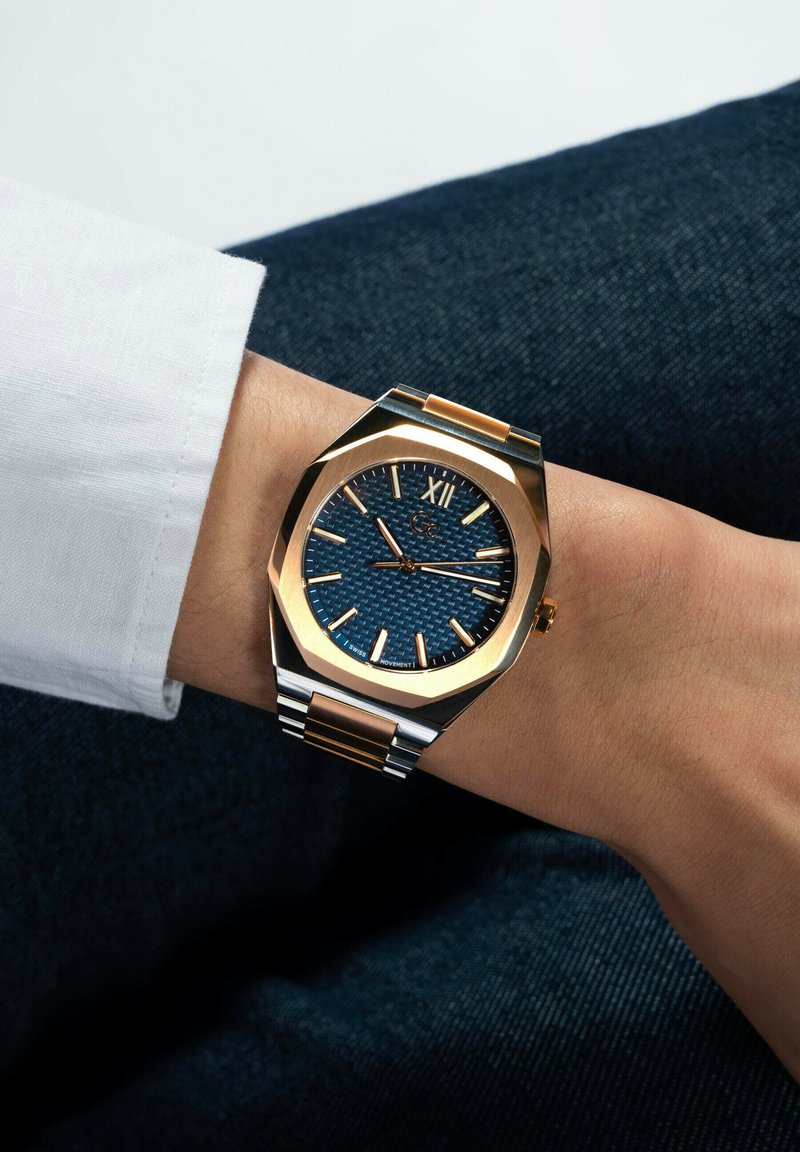 Auksinis ir sidabrinis riešo laikrodis su mėlynu tekstūruotu ciferblatu ir auksiniais valandų žymekliais, uždėtas ant riešo, apkaltinant tamsiai mėlynas džinsus ir baltą marškinių rankovę.