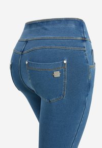 Primer plano de jeggings de mezclilla azul con cintura alta y bolsillo trasero que presenta una pequeña placa metálica con logo en el lado derecho.