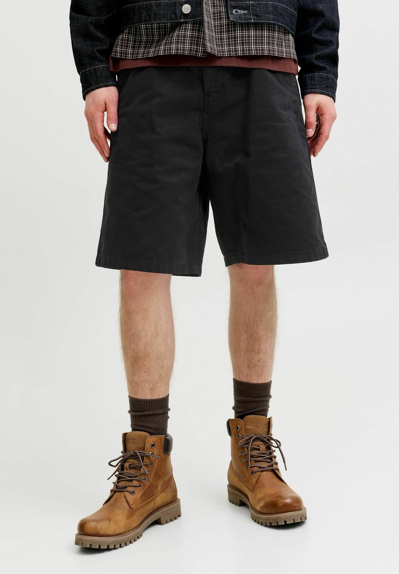 Homme portant un short noir au niveau des genoux, des chaussettes marron et des bottes en cuir beige à lacets, debout devant un fond clair uni.