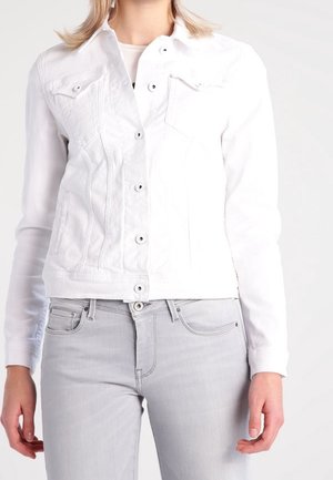 Kobieta w białej kurtce dżinsowej na białym topie, w połączeniu z jasnoszarymi dopasowanymi jeansami, stojąca na jednolitym tle.
