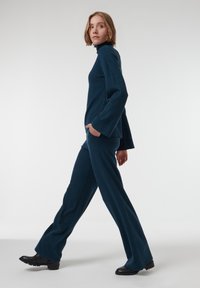 Pull en tricot teal foncé avec un col haut et des manches larges, associé à un pantalon évasé. Le mannequin se tient dans un cadre neutre, tourné vers la gauche.