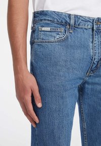 Kék farmernadrág, slim fit szabású, klasszikus öt zsebes dizájnnal, hátul kis "Guess Jeans" címkével.