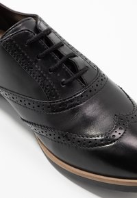 Scarpa da dress nera in pelle con dettagli brogue, caratterizzata da una finitura liscia, chiusura con lacci e una suola marrone leggermente rialzata.