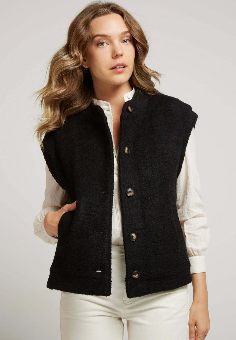 Gilet en polaire noir avec un col, doté de gros boutons et de deux poches latérales. Porté par-dessus une chemise de couleur claire, sur fond neutre.