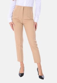 Pantaloni beige a vita alta con tasche laterali, texture liscia e gamba affusolata, abbinati a décolleté nere a punta, con una camicia bianca impeccabile.