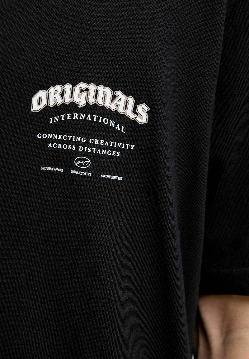 Sort bomulds t-shirt med et stort hvidt og creme farvet grafisk print med teksten: "ORIGINALS INTERNATIONAL CONNECTING CREATIVITY ACROSS DISTANCES."