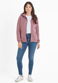 Light mauve zip-up hoodie with a drawstring hood, kangaroo pockets, and ribbed cuffs, paired with blue skinny jeans and white sneakers.