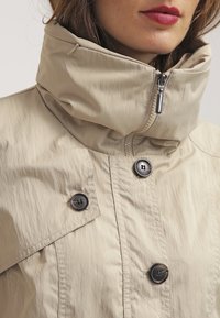 Chaqueta ligera beige con un alto cuello, cremallera frontal y dos botones negros como acentos. Presenta una textura arrugada y líneas limpias.