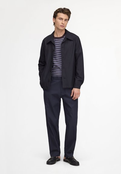 Jeune homme debout les mains dans les poches, portant une veste noire, une chemise rayée, un pantalon sombre et des mocassins noirs sur fond blanc.