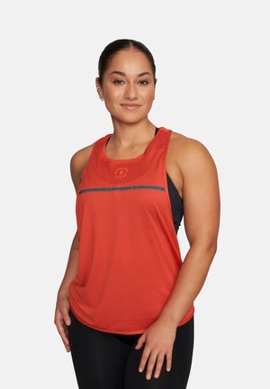 Kobieta ubrana w czerwony sportowy top bez rękawów i czarne legginsy, stojąca pewnie na tle jasnego, jednolitego tła.