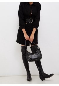 Borsa a mano in pelle nera con texture a coccodrillo, borchie argentate e un dettaglio a nappa. Tenuta da una persona che indossa un vestito nero e stivali alti fino al ginocchio.