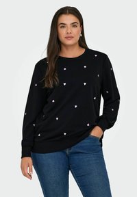 Schwarzes Sweatshirt aus weichem Material, mit verstreuten rosa Herzmustern. Lange Ärmel mit gerippten Bündchen. Kombiniert mit blauen Jeans.
