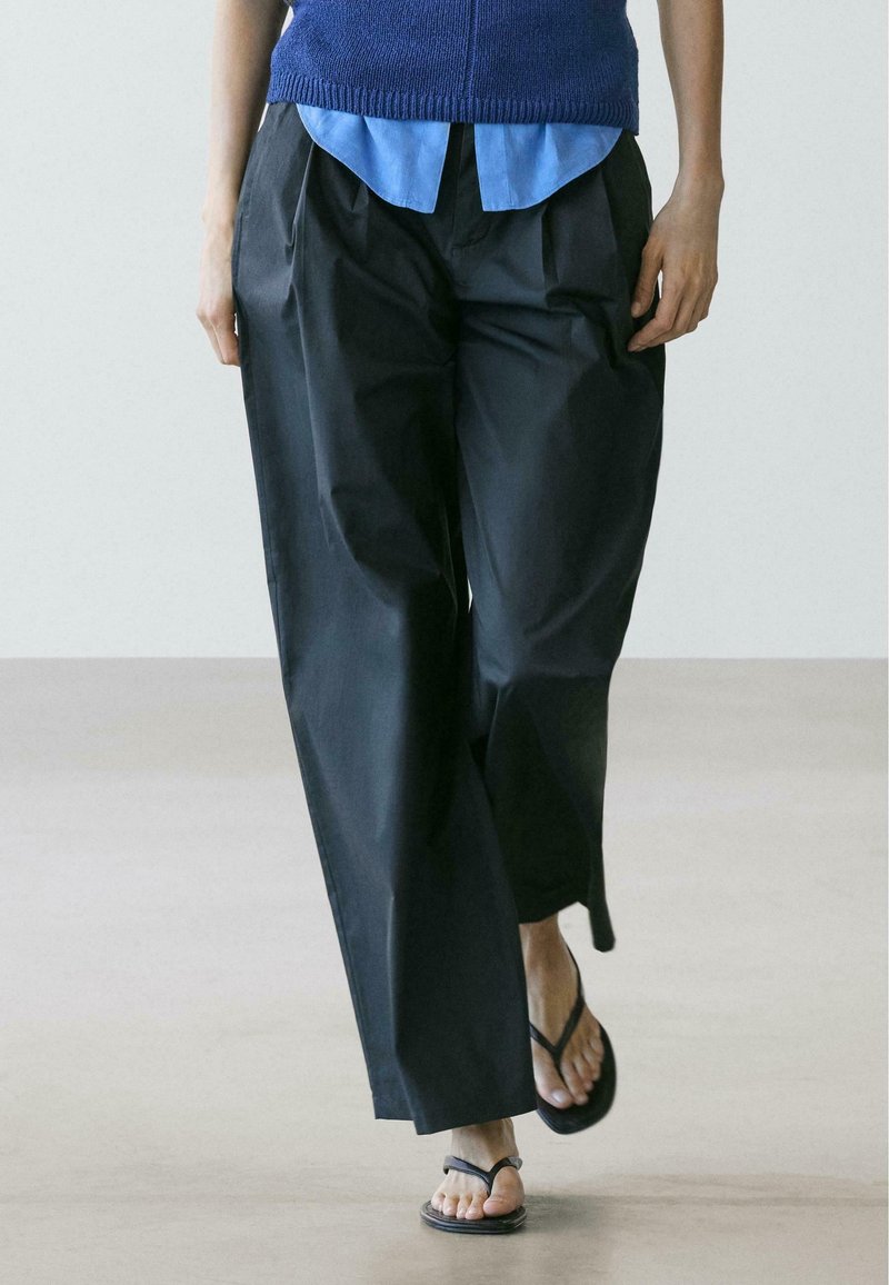 Personne marchant portant un pantalon noir à jambes larges, une chemise bleue, un pull bleu et des sandales noires en tongs sur un sol lisse et clair.