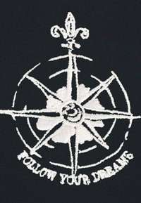 Designo di una bussola ricamata in bianco su un tessuto nero, con un fleur-de-lis e il testo "SEGUITA I TUOI SOGNI" in basso.