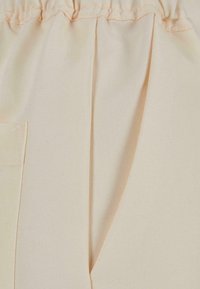 Pantalon en tissu beige avec ceinture élastique, pli devant et poche sur la couture latérale visible.