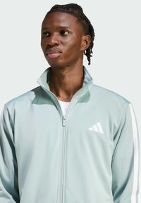 Svijetlozelena jakna na zip s visokim ovratnikom, bijelim ukrasnim prugama i logom na lijevoj strani prsnog koša, izrađena od glatkog, sportski podesnog materijala.