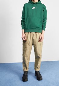 Grön bomullshoodie med framficka och vitt logotyp, kombinerad med beige cargobyxor med sidfickor och svarta snörstövlar.
