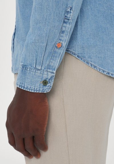 Light blue denim shirt with a cuff showing orange and green buttons. Hand resting on light beige fabric, highlighting texture contrast.