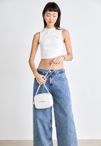 Balts īsais bez piedurknēm tops ar augstu kakla izgriezumu, saskaņots ar brīviem zilos džinsos. Mazs balts crossbody soma ar "Tommy Jeans" logo.
