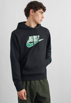 Młody mężczyzna w czarnej bluzie z kapturem Nike z zielonym logo, z rękami w kieszeniach, patrzący na bok, ubrany w oliwkowe spodnie na jednolitym tle.