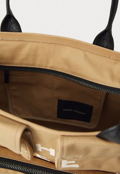 Lähikuva vaaleanruskean kankaisen laukun sisätilasta, jossa on musta vetoketju, mustat kahvat, sisätaskut ja Marc Jacobs -logo ommeltuna sisälle.