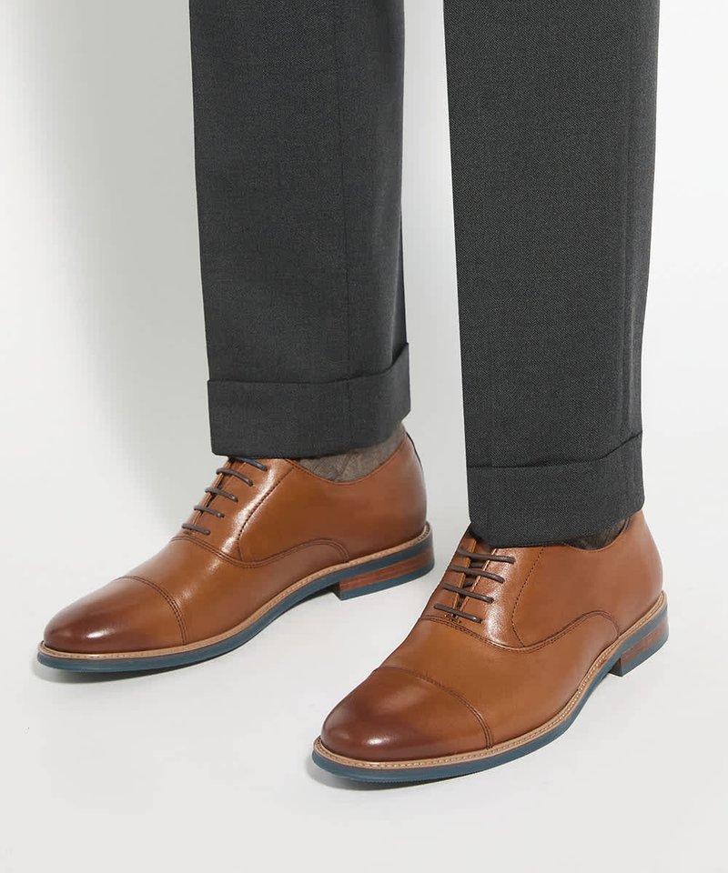 Chaussures habillées en cuir marron avec un laçage fermé portées avec un pantalon gris foncé à revers sur fond blanc.