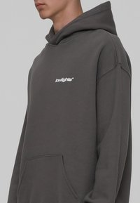 Donkergrijze hoodie met een voorzak en een capuchon met trekkoord. Bevat witte logo tekst op de borst. Zachte, katoenmix stof.