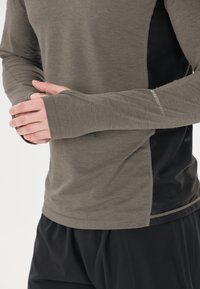 Pitkähihaista paitaa oliivinvihreänä, jossa on pehmeä rakenne, tiukka istuvuus ja mustat verkkopaneelit sivuilla hengittävyyden ja liikkuvuuden parantamiseksi.