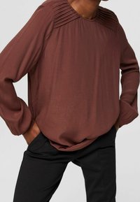 Blusa marrone a maniche lunghe con dettagli plissettati sulle spalle, caratterizzata da una vestibilità comoda e una texture liscia, abbinata a pantaloni neri.