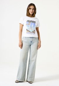 Camiseta gráfica blanca con diseño de texto colorido y conchas, combinada con pantalones anchos de mezclilla azul claro y zapatos metálicos.