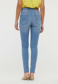 Jean en denim bleu skinny avec une taille haute, dotés de poches arrière et de coutures jaunes. Le modèle porte un top jaune et des talons beiges.