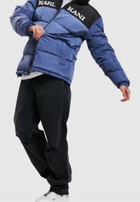 Blå puffjacka med en svart axelpanel, med vit logotyp. Kombinerad med svarta urblekta jeans och vita sneakers.