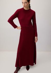 Sötétvörös kötött ruha, illeszkedő felső résszel, hosszú ujjakkal és redős szoknyával. Az anyag ribbelt strukturával rendelkezik.