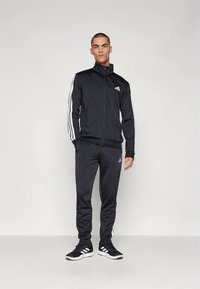 Čierny teplákový komplet z ľahkého materiálu s bielymi pruhmi pozdĺž rukávov a bokov, obsahujúci bundu na zips a zodpovedajúce nohavice.