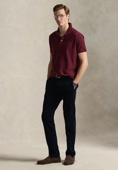 Burgunderfarget poloshirt med korte ermer, kombinert med mørkeblå bukser og brune mokkasin-sko. Tilbehør med runde briller og klokke.