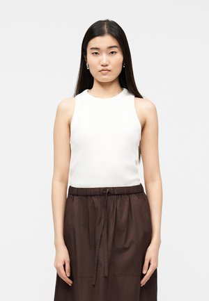 Giovane donna con lunghi capelli scuri indossa un top bianco senza maniche e una gonna marrone con coulisse, in piedi davanti a uno sfondo semplice.