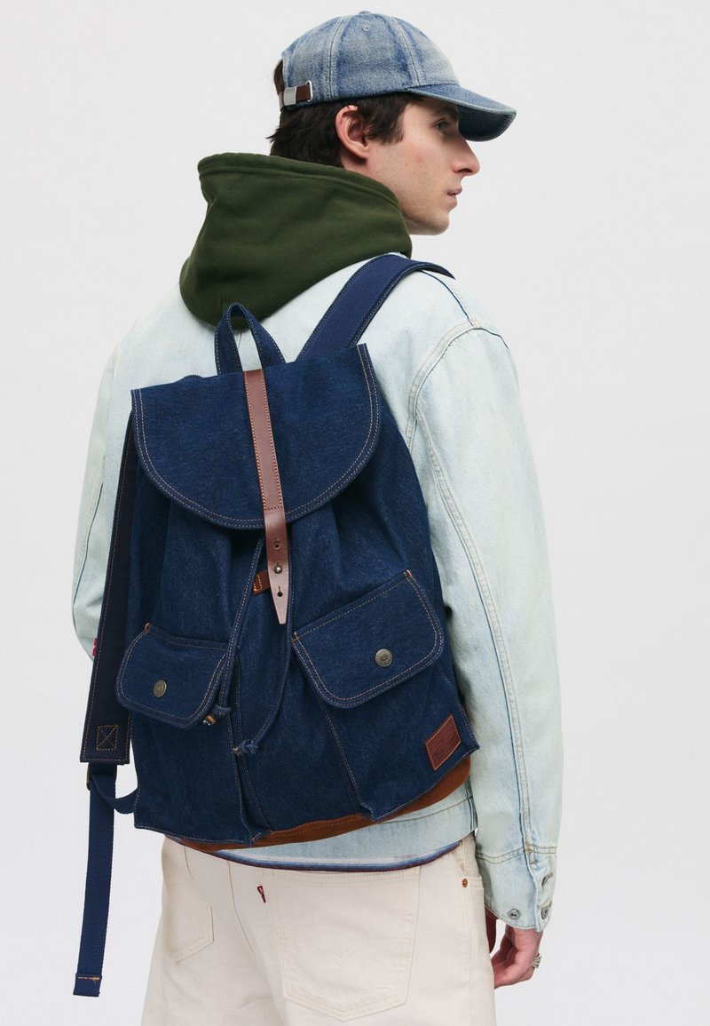 Zaino in denim di colore blu navy, con dettagli in pelle, due tasche frontali, una patta pieghevole e spallacci regolabili.