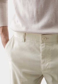 Helle beigefarbene Baumwollhose im Slim-Fit mit einem Frontknopfverschluss und seitlichen Taschen, kombiniert mit einem hellgrauen Strickpullover.