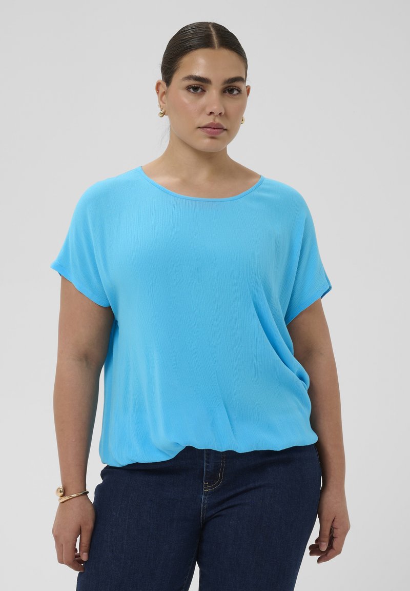 Femme aux cheveux foncés portant un haut bleu à manches courtes et un jean foncé, debout devant un fond clair uni, portant des boucles d'oreilles et un bracelet en or.