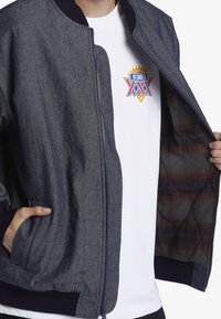 Denim bombarjacka med marinblå ribbade muddar och nederkant, dragkedja och rutigt foder. Bärs över en vit grafisk t-shirt.