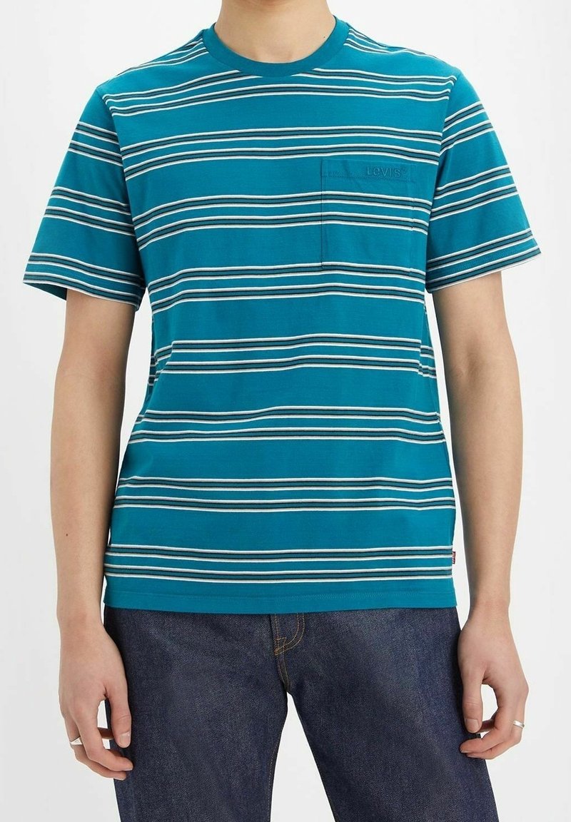 Homme portant un t-shirt à manches courtes col rond couleur bleu sarcelle avec des rayures horizontales blanches et noires, et un jean bleu foncé, debout les bras détendus.
