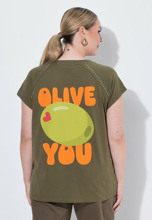 Donna con coda di cavallo bionda che indossa una maglietta verde oliva con una grande oliva in stile cartone animato e il testo "Olive You" in arancione sulla schiena.