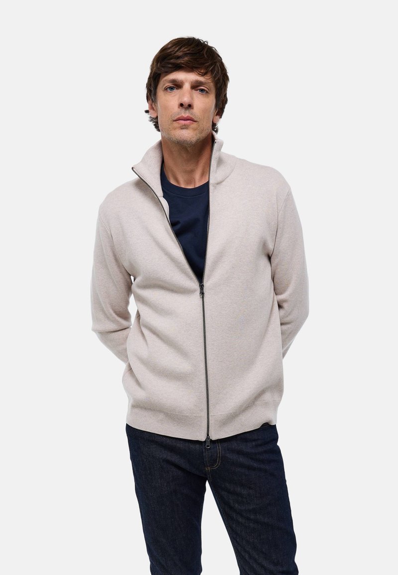 Gilet zippé beige clair avec col montant, manches et ourlet côtelés, porté sur un t-shirt bleu marine, associé à un jean bleu foncé.