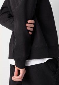 Asmuo dėvi juodą ribotą džemperį ir baltą marškinėlį, viena ranka laiko priešingo rankovės galą už nugaros.