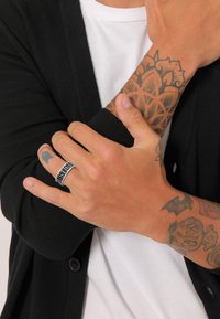 Inel din metal argintiu cu inscripția "JUSTICE", purtat pe o mână cu tatuaje vizibile, pe un fundal format dintr-un cardigan negru și o cămașă albă.