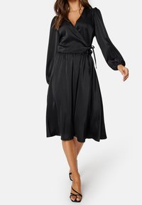 Femme portant une robe portefeuille en satin noir avec de longues manches bouffantes et des talons noirs à lanières, marchant sur un fond blanc.