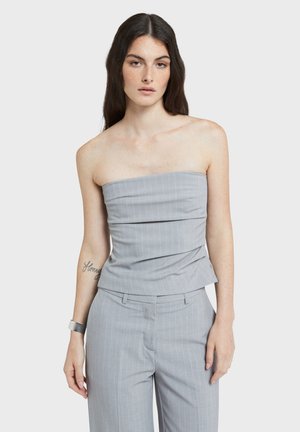 Femme aux longs cheveux noirs portant un haut gris rayé sans bretelles et un pantalon assorti, posant devant un fond blanc uni.